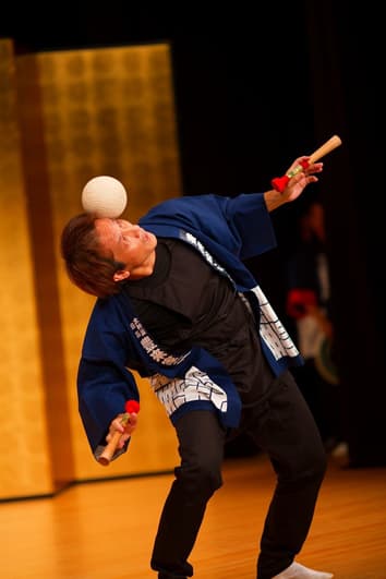 バチを両手に持ちながら鞠を側頭部に載せる様子