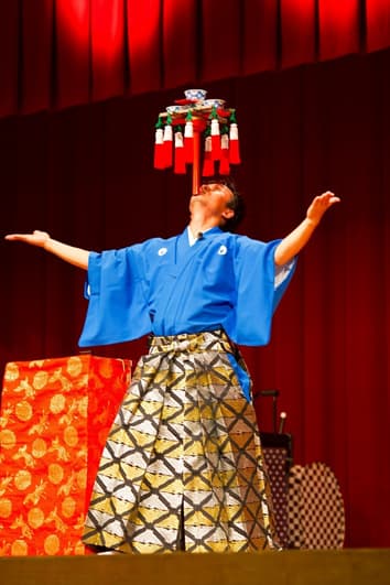 あごの上に立てたバチにたくさんの茶碗や房がのっている様子